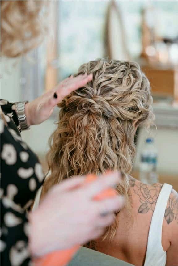 Stylist arranging intricate braided hairstyle on woman with curly blonde hair and shoulder tattoo.
