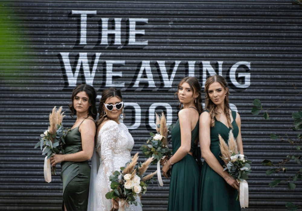 Bride and bridesmaids in green dresses pose with flowers outside "The Weaving Room."