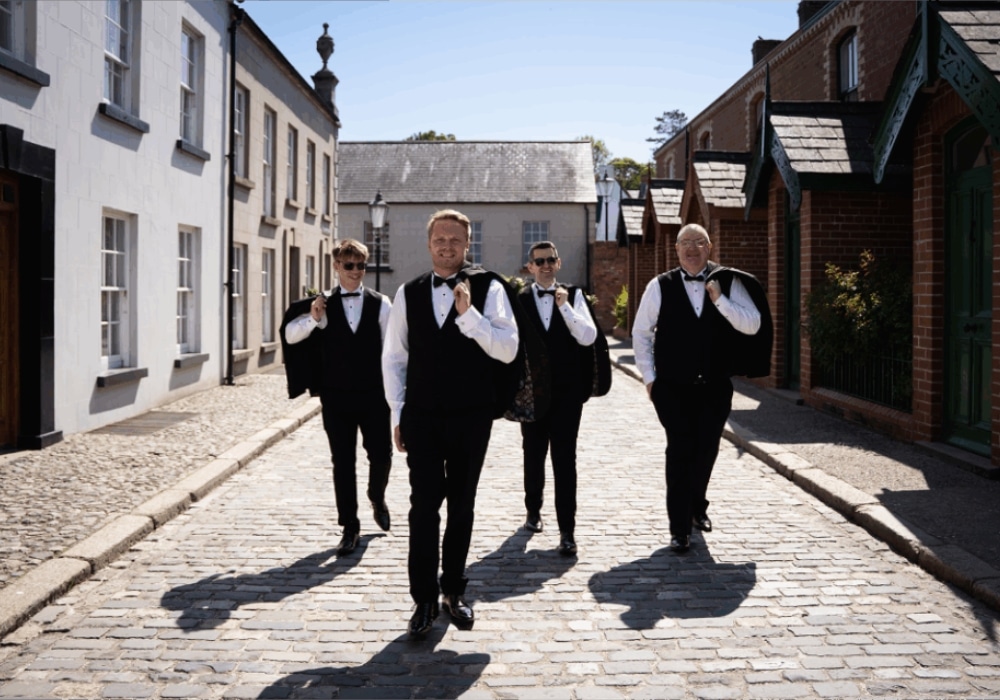 Four men in formal attire walk confidently down a cobblestone street.