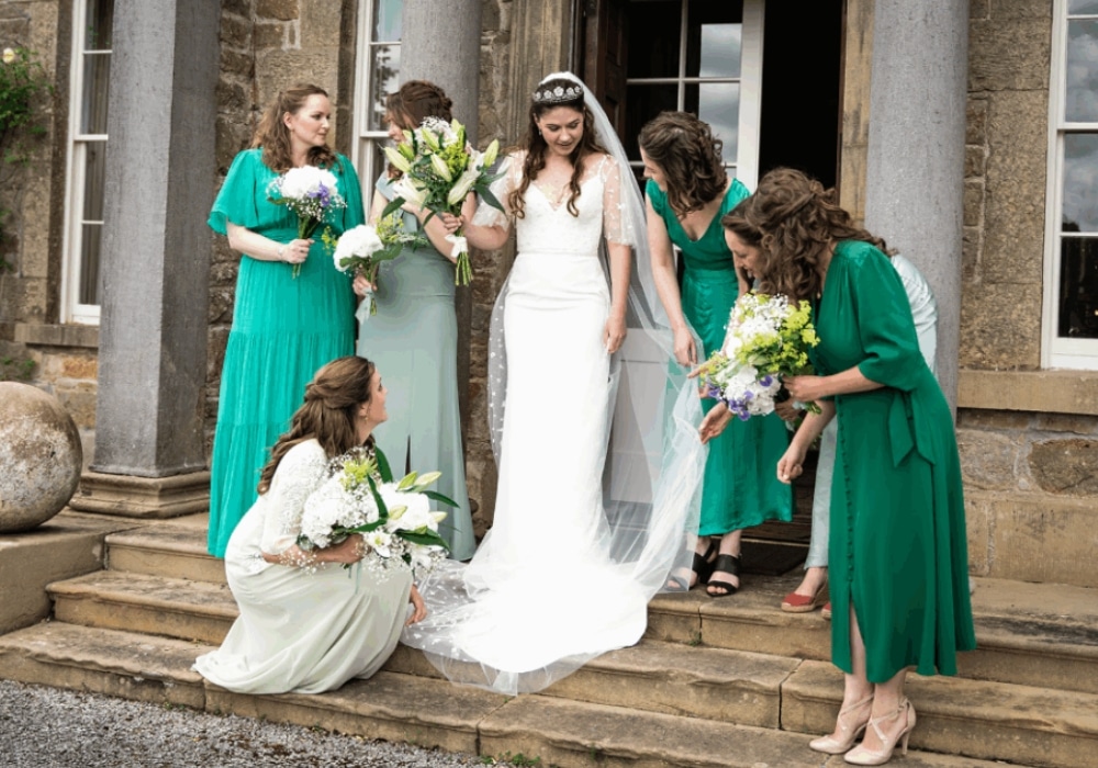 Bride with bridesmaids in green dresses holding bouquets on stone steps.