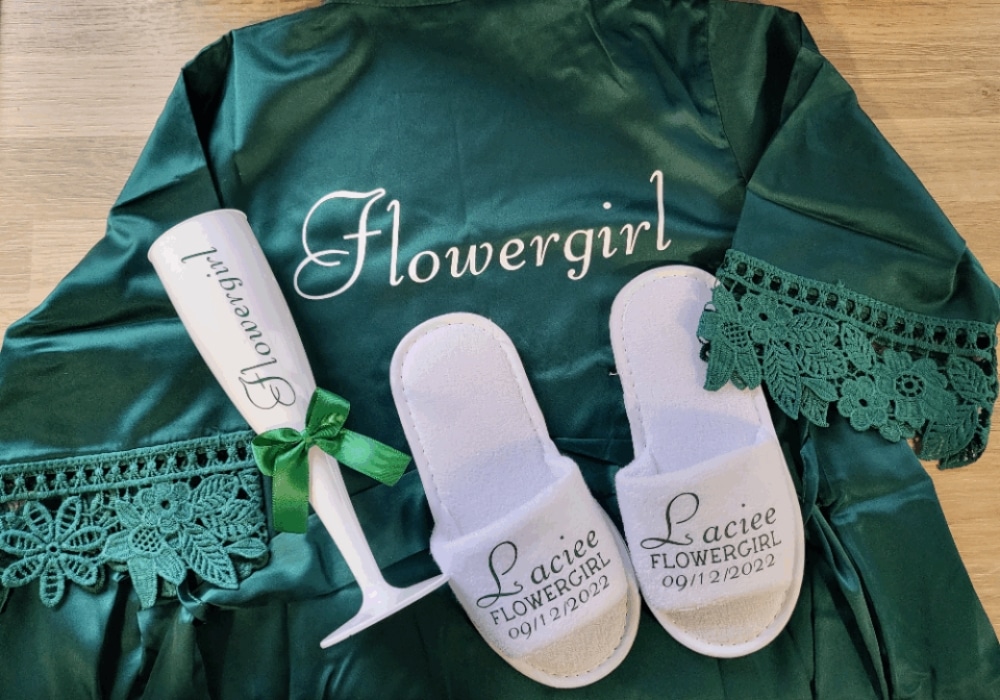 Green flower girl robe, white slippers, and engraved champagne flute with green ribbon on a wooden surface.
