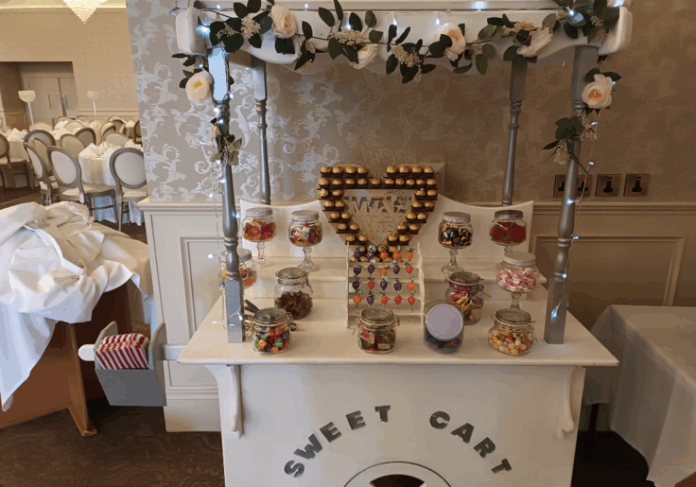 A sweet cart decorated with flowers, displaying assorted candies and chocolate jars at an event venue.