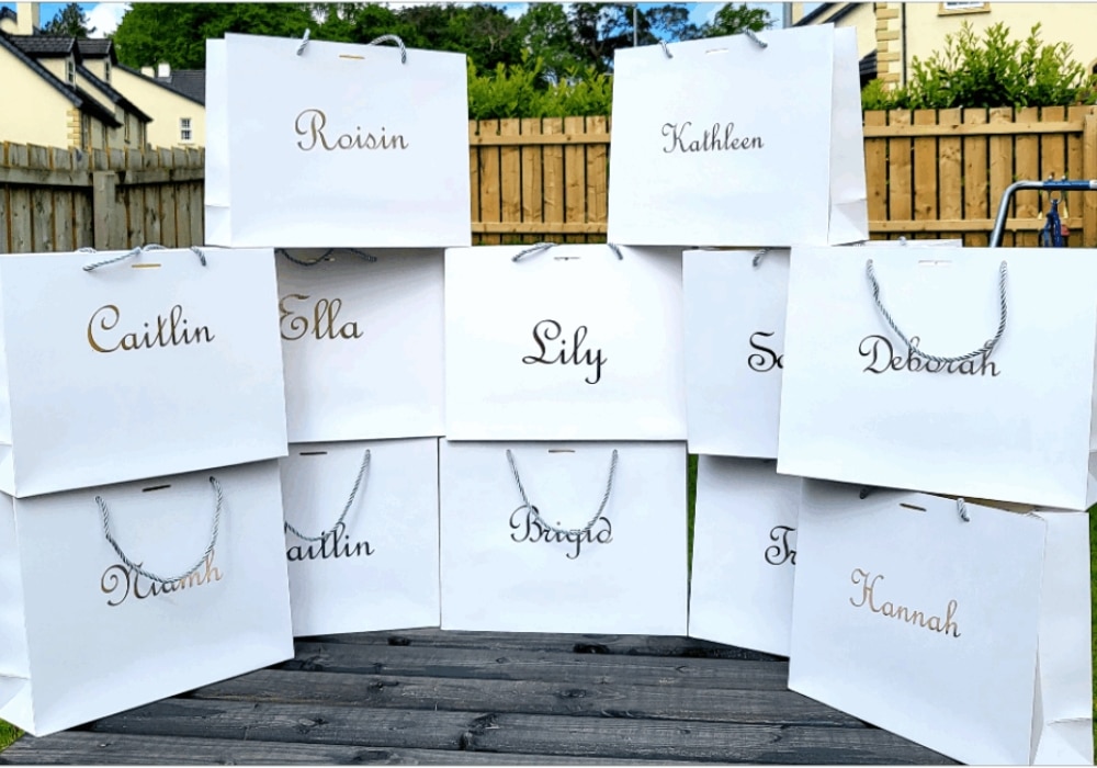 White gift bags with names arranged outdoors on a wooden surface.