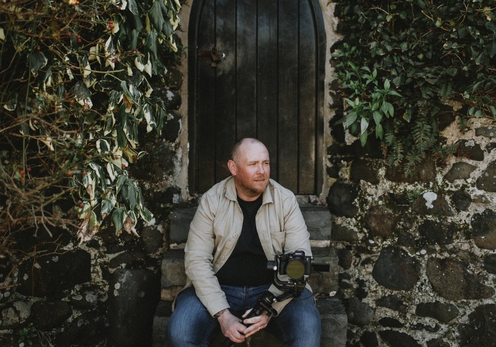 Man with a camera sitting on stone steps by a rustic wooden door surrounded by ivy and stone wall.