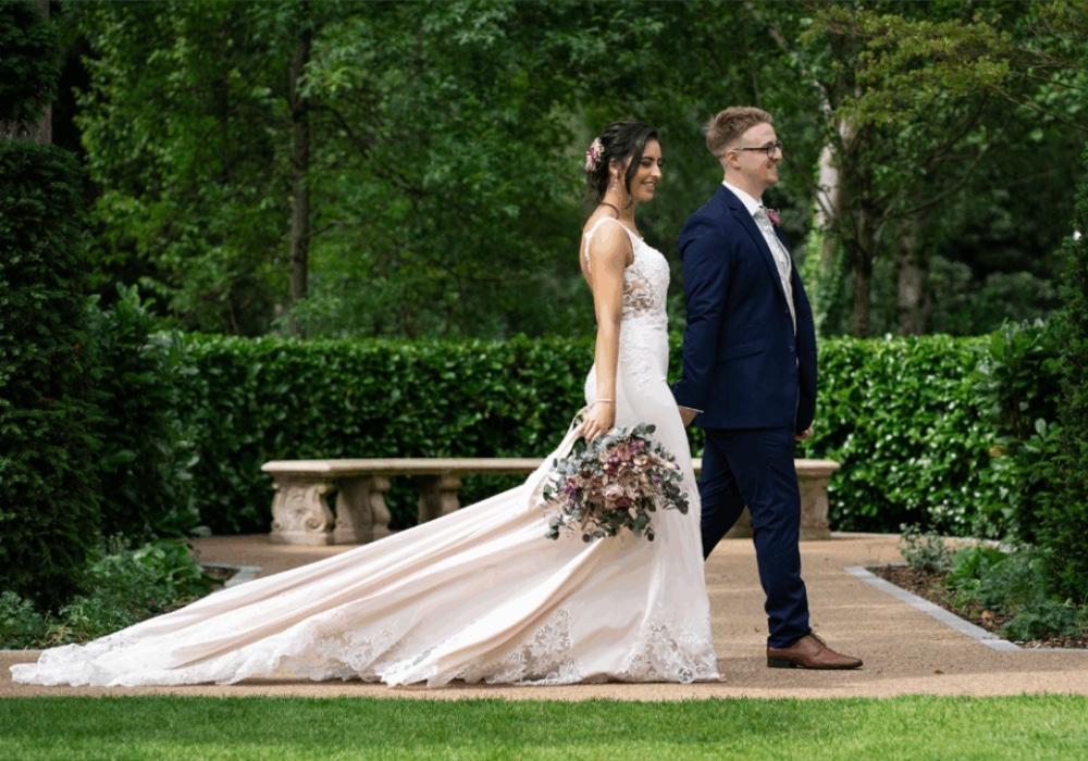 Bride and groom walking in a garden, bride holding a bouquet, wearing a white gown, groom in a navy suit.