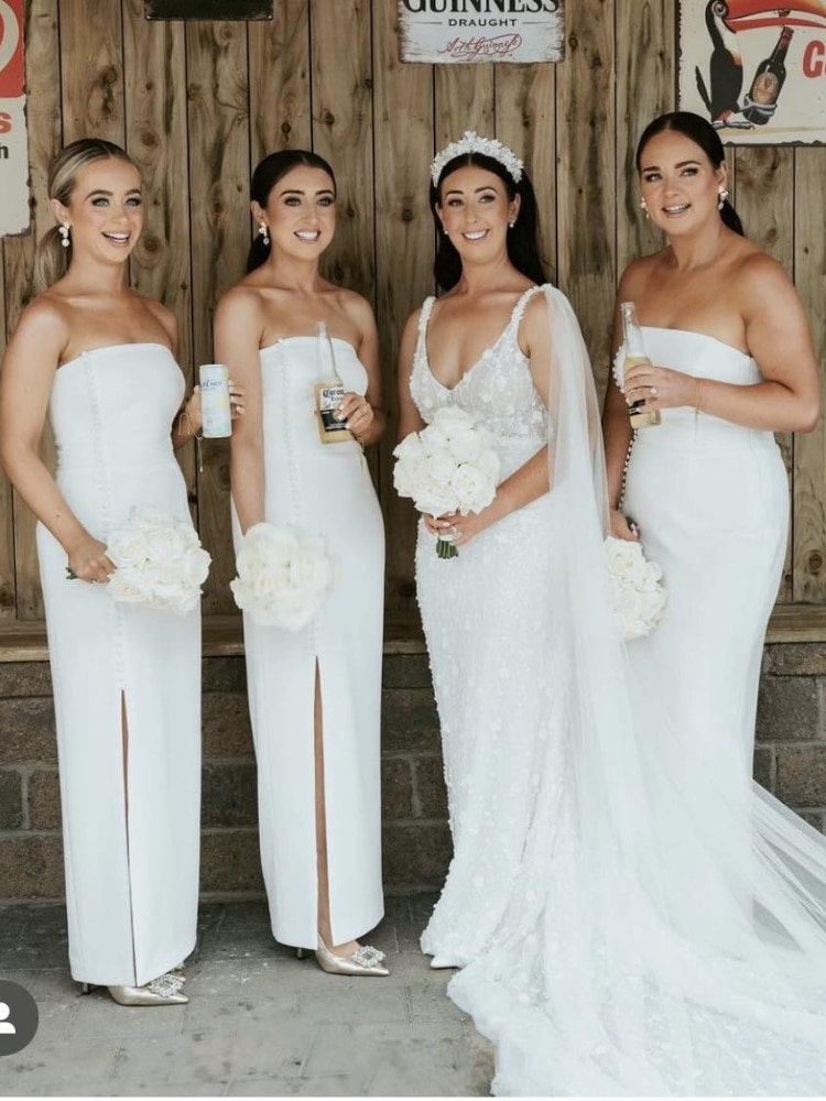 Bridesmaids and bride in white dresses holding bouquets and drinks.