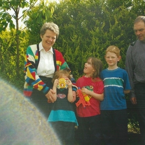 A group of people, including children, smiling outdoors in front of greenery; one child holds a toy camera.