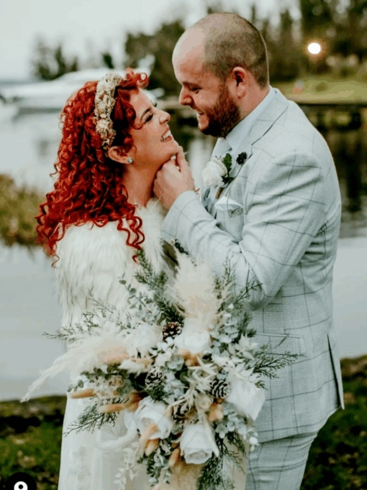 Bride and groom embracing near a lake, holding a bouquet with greenery and white flowers.