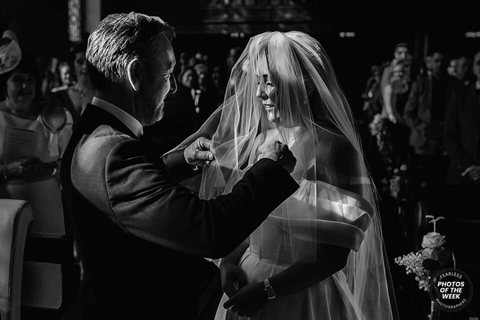 Father lifting bride's veil at wedding ceremony, surrounded by guests in a dimly lit venue, black and white photo.