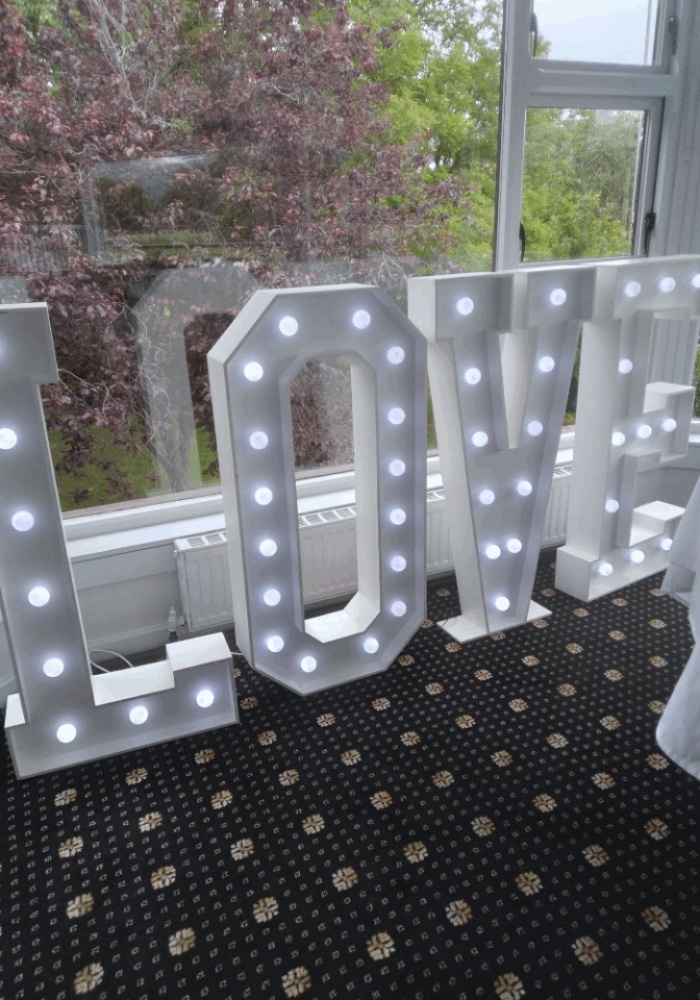 Illuminated "LOVE" sign with white lights, placed indoors near a window with greenery in the background.