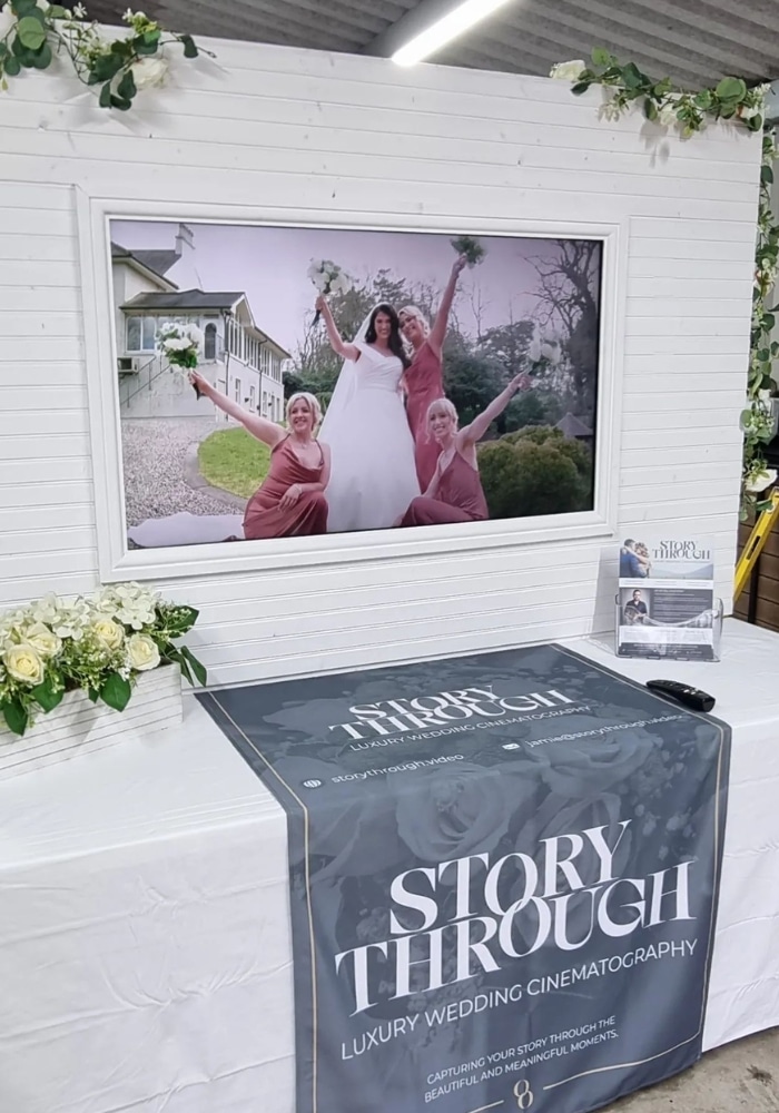 Wedding cinematography display with a framed photo of a bride and bridesmaids on a white wall, flowers, and a promotional banner.