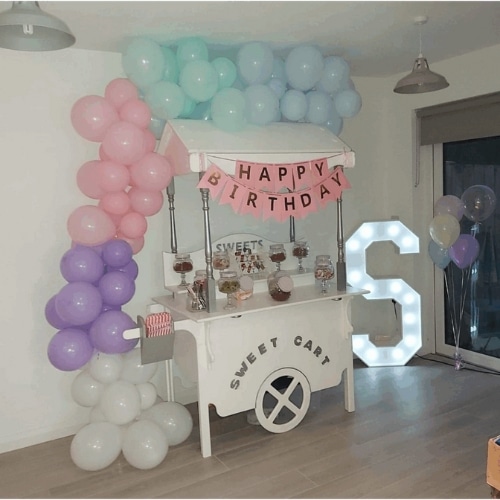 Sweet cart with a pastel balloon arch, "Happy Birthday" bunting, and illuminated letter "S" in a room.