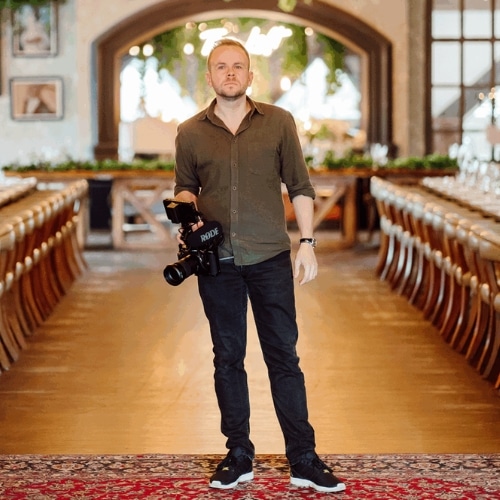 Man holding camera stands in decorative banquet hall with long dining tables and archway.