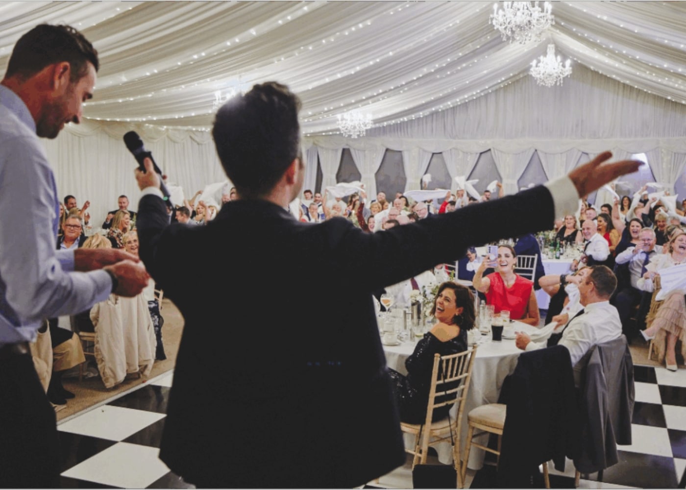 Wedding reception with guests cheering under a decorated white tent, a person with a microphone in the foreground.