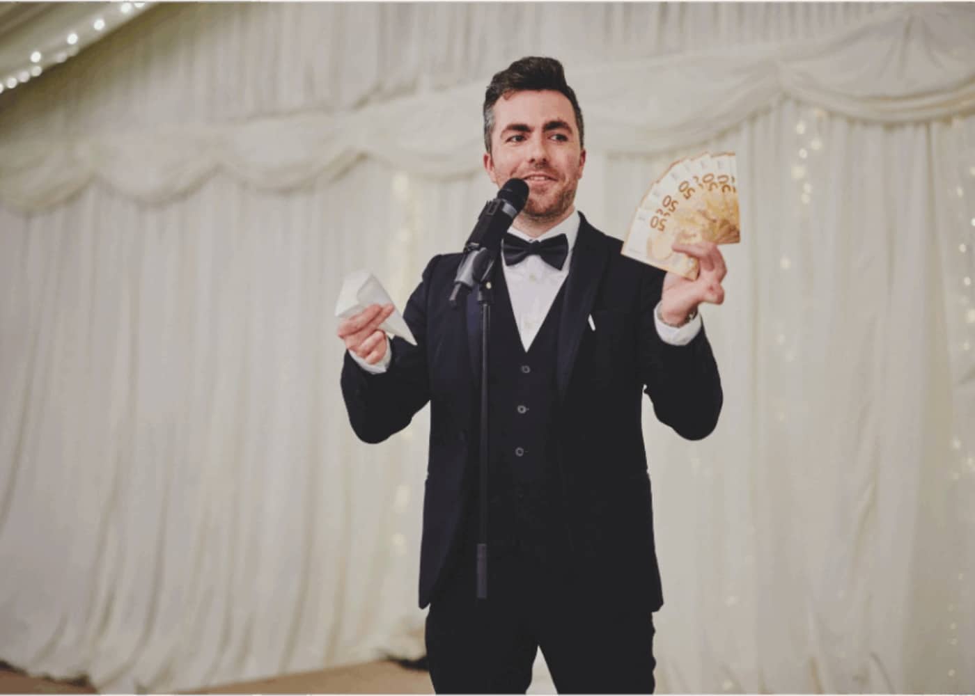 Man in a suit holding a microphone and money in a decorated event setting.