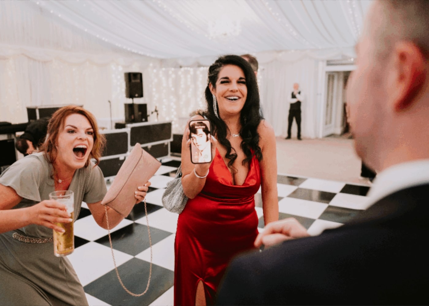 Two women in an elegant, festive setting, one holding a phone with a photo, the other laughing with a drink.