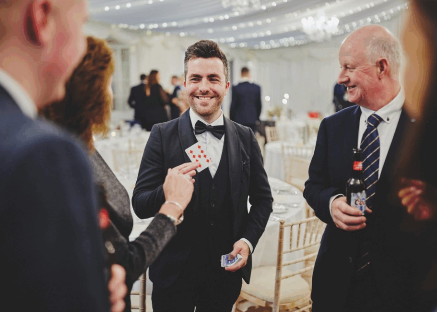 Magician performing a card trick at an event, smiling with attendees around, in a decorated venue with string lights.
