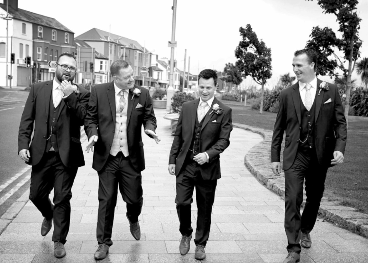 Four men in suits, smiling and walking on a sidewalk in a city, black and white photo.