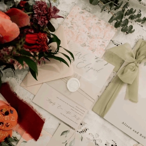 Wedding stationery with floral designs and ribbon, including an elopement announcement, surrounded by flowers and greenery.