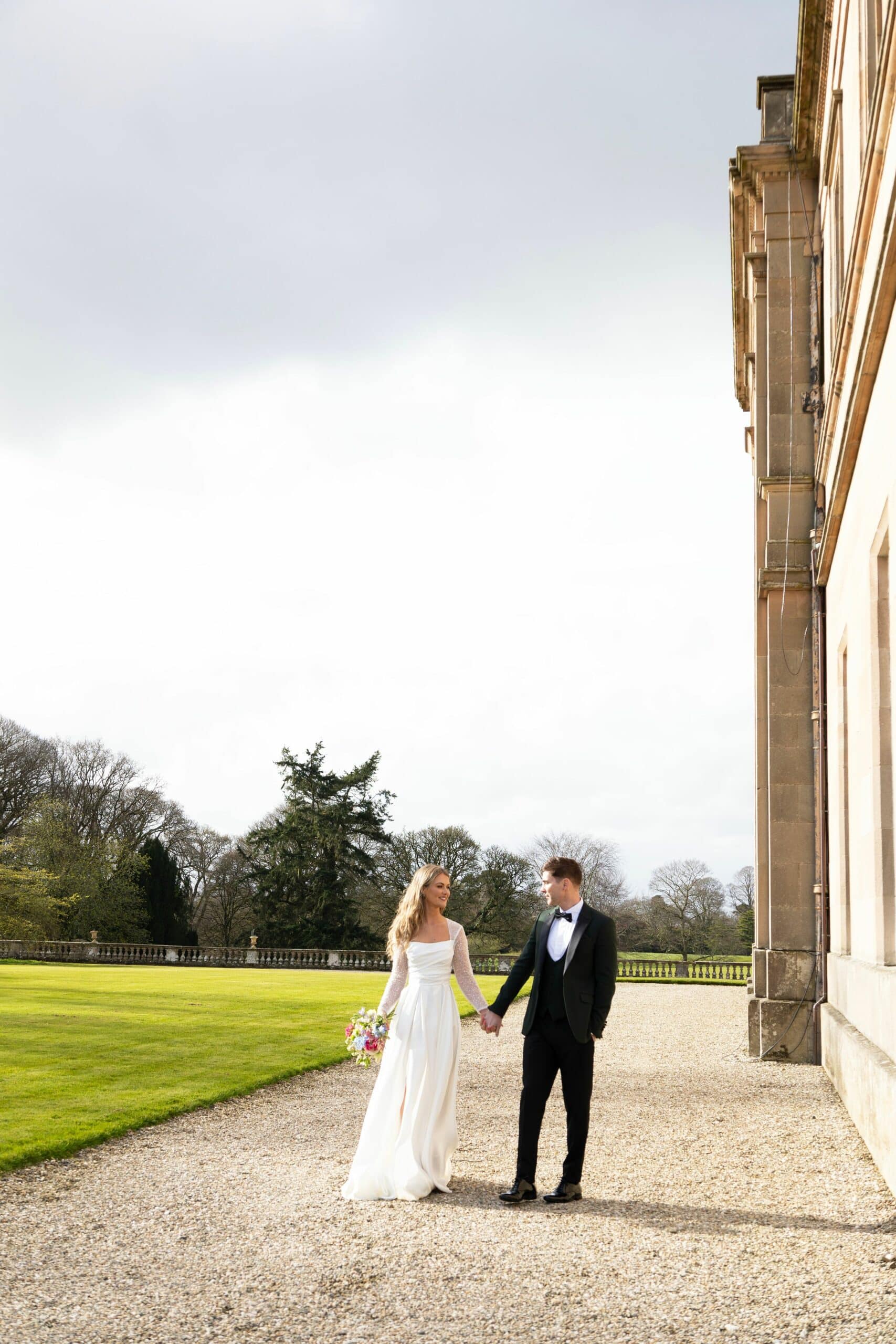 Bride and groom holding hands, walking on a gravel path beside a historic building, surrounded by a green lawn and trees.