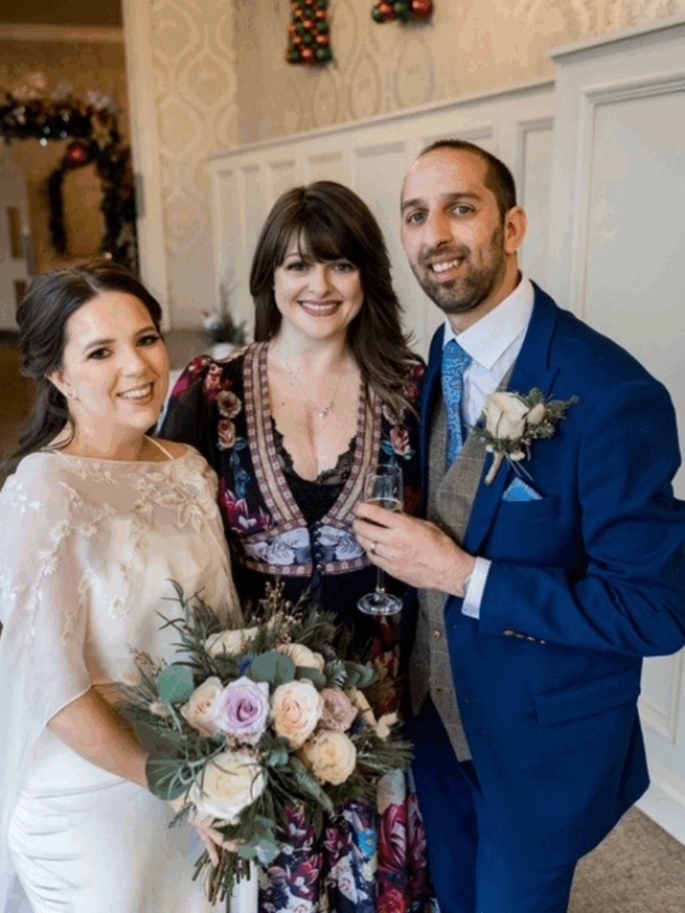 Bride and groom posing with a friend, indoors, holding a bouquet and a glass of champagne.