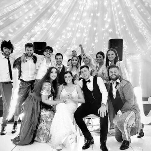 Wedding party posing under string lights in a decorated venue, with the couple seated at the front, smiling guests around them.