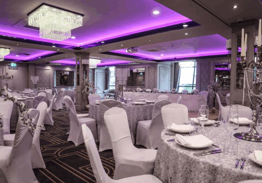 Elegant banquet hall with round tables, white chairs, crystal chandeliers, and purple lighting.