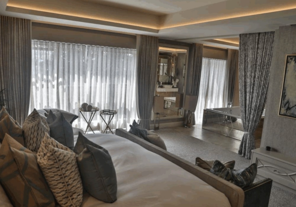 Luxury bedroom with large windows, elegant curtains, and modern decor, featuring a bed with decorative pillows and a spacious view.