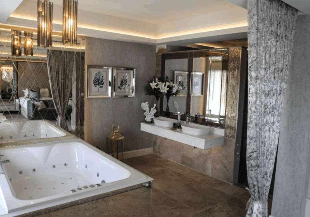 Luxury bathroom with a large bathtub, dual sinks, elegant lighting, and decor, featuring mirrored walls and tile flooring.