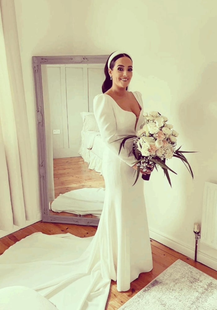 Bride in an elegant white gown holding a bouquet, standing in a bright room with a large mirror.
