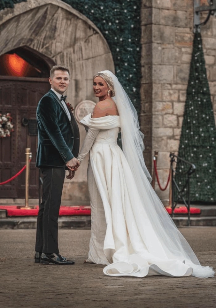 Bride and groom holding hands outside a stone building, wearing wedding attire and smiling at the camera.