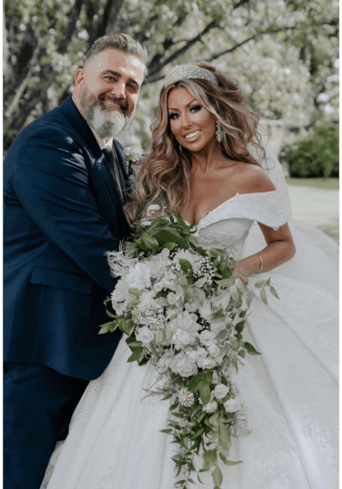 Bride and groom smiling, holding a lush bouquet with white flowers and greenery, in an outdoor setting.