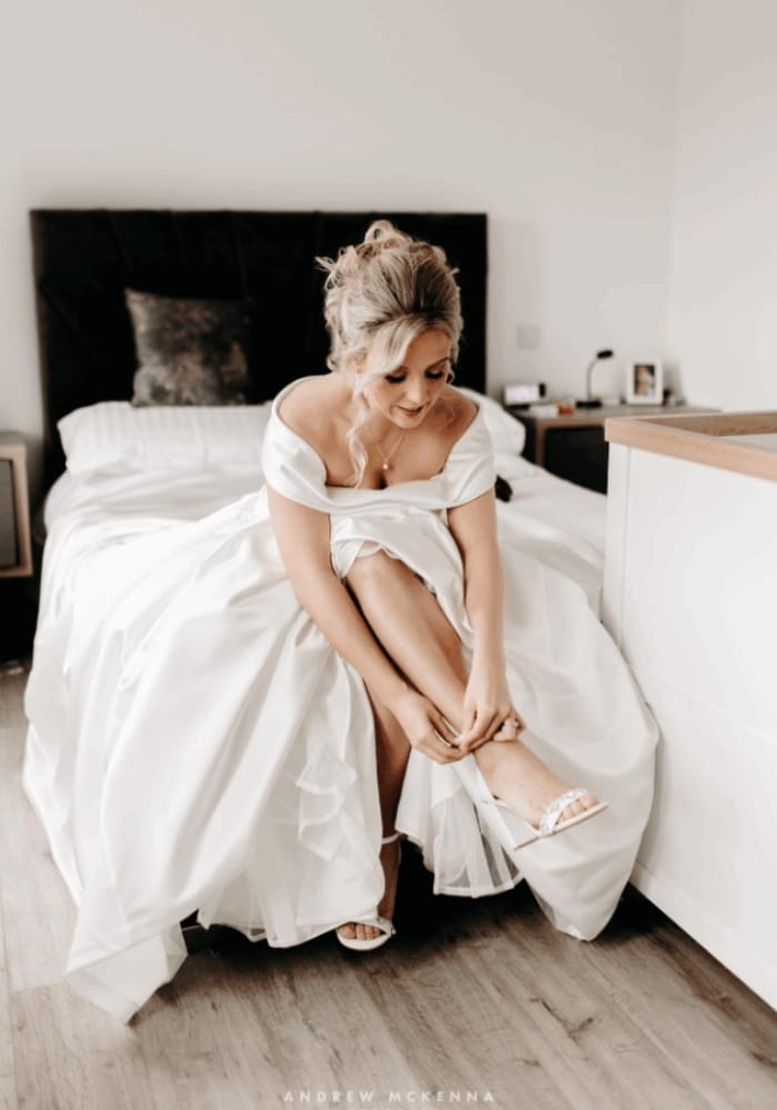 Bride sitting on a bed, adjusting her white shoe while wearing a wedding dress.