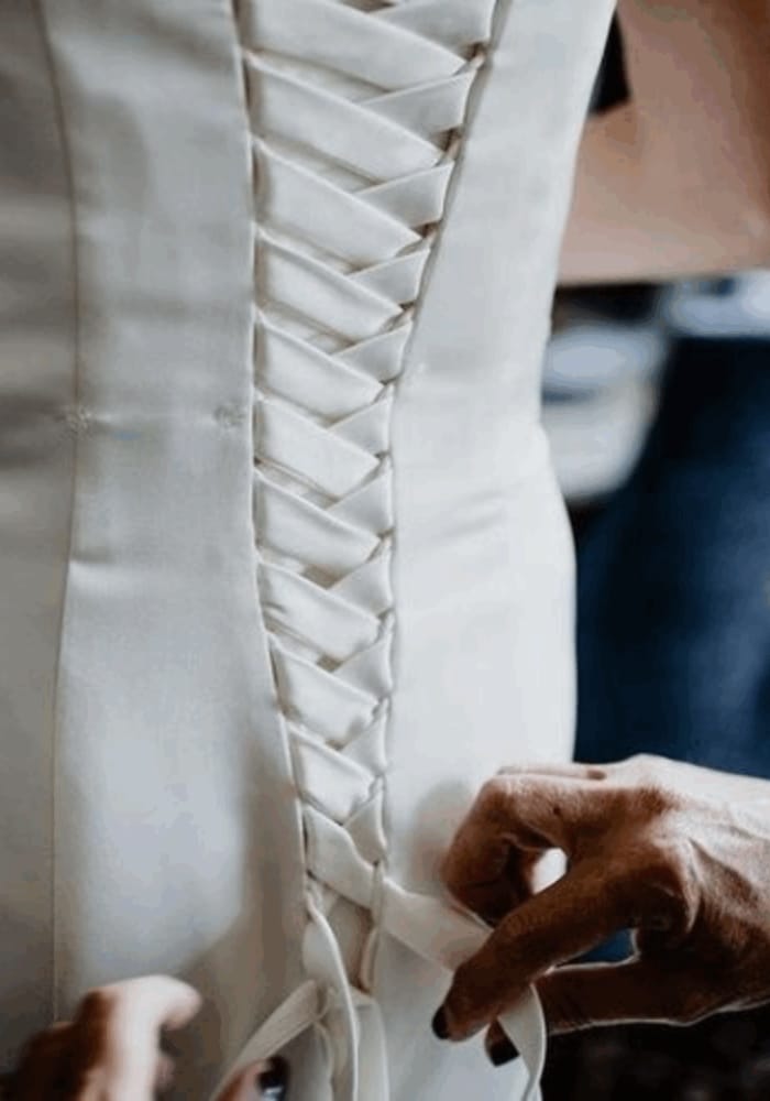 Hands lacing up the back of a white corset-style dress.