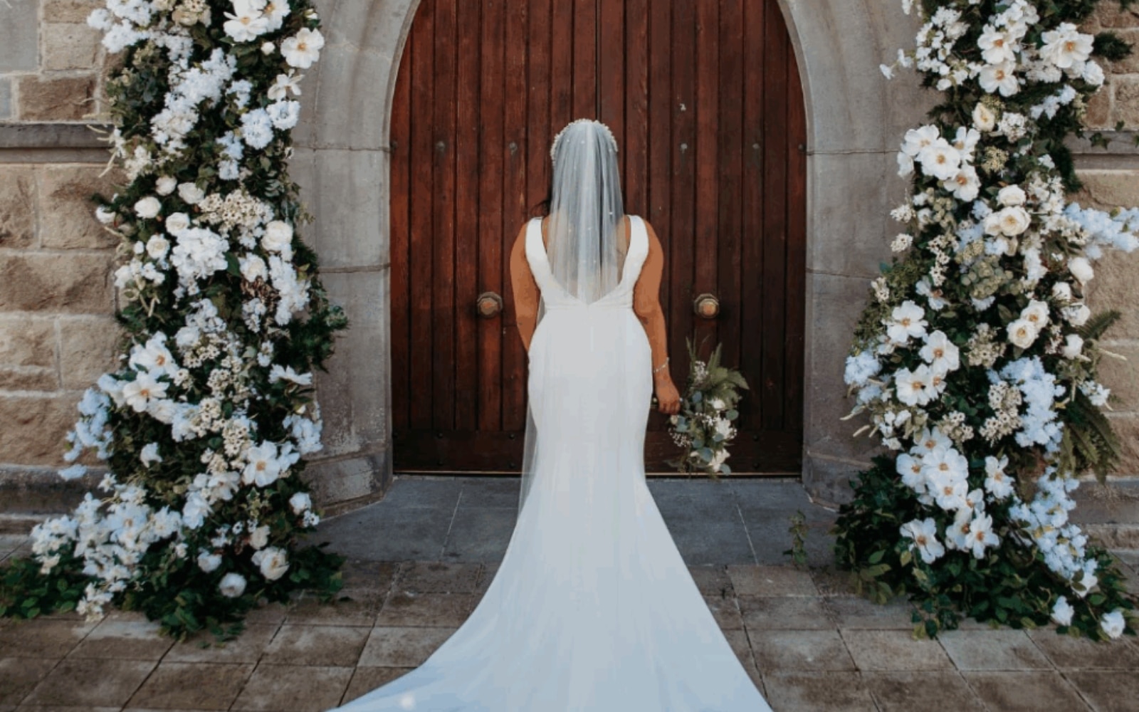 Bride in white gown with bouquet, facing ornate wooden door adorned with floral arrangements.