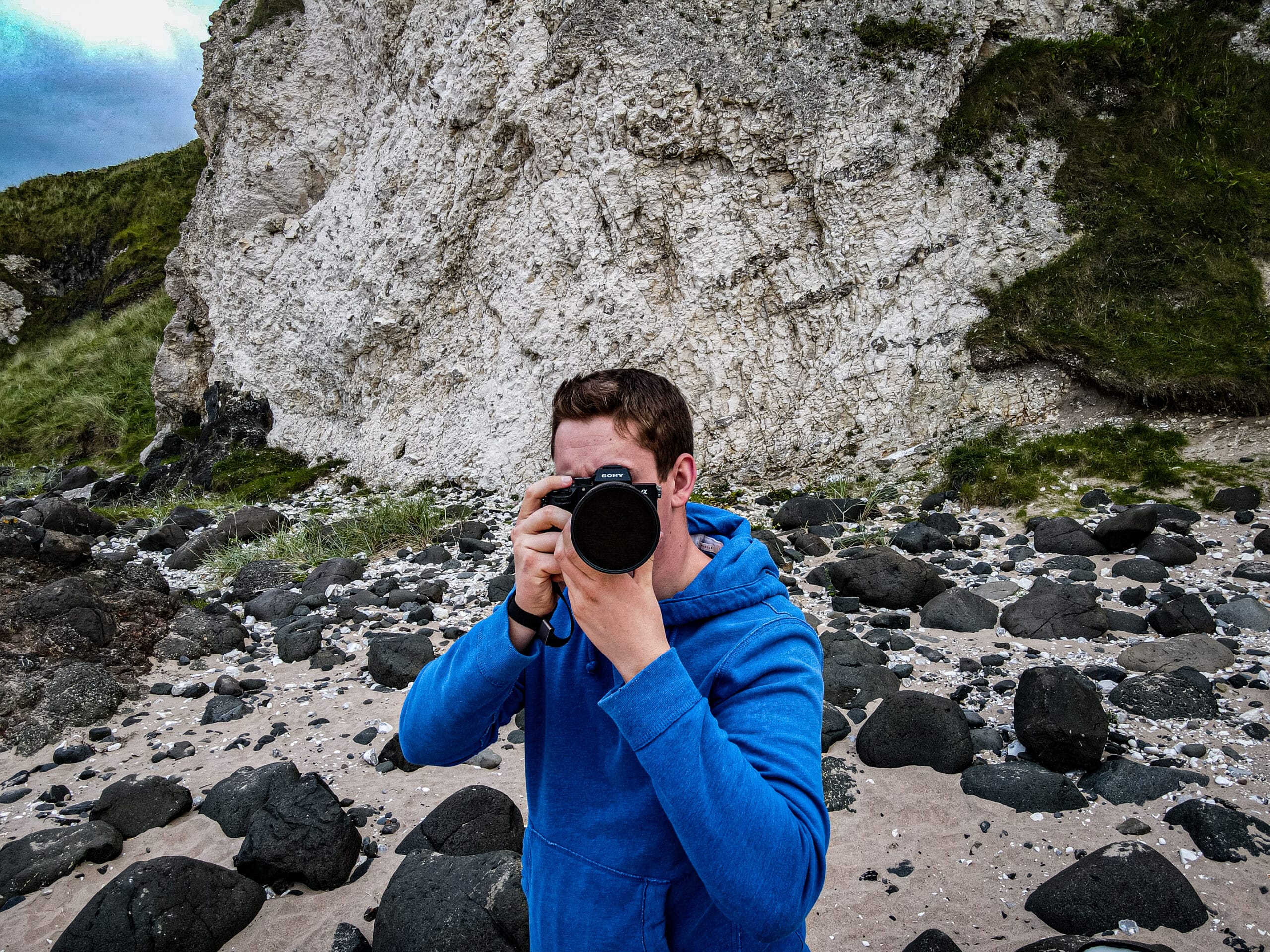 A person in a blue hoodie taking a photograph on a rocky beach with white cliffs in the background.