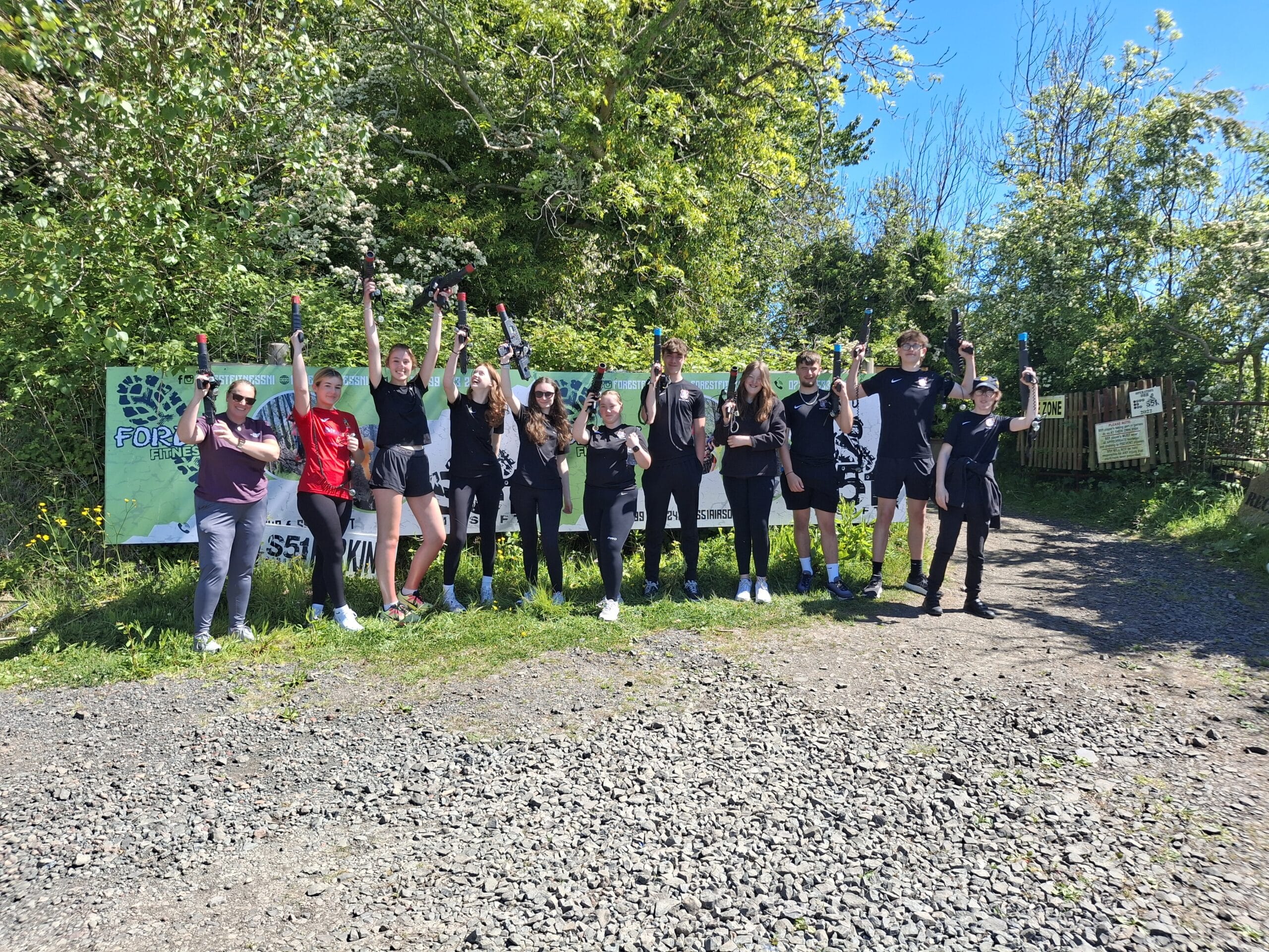 Group of people holding laser tag guns outdoors in front of a "Forest Fitness" banner on a sunny day.