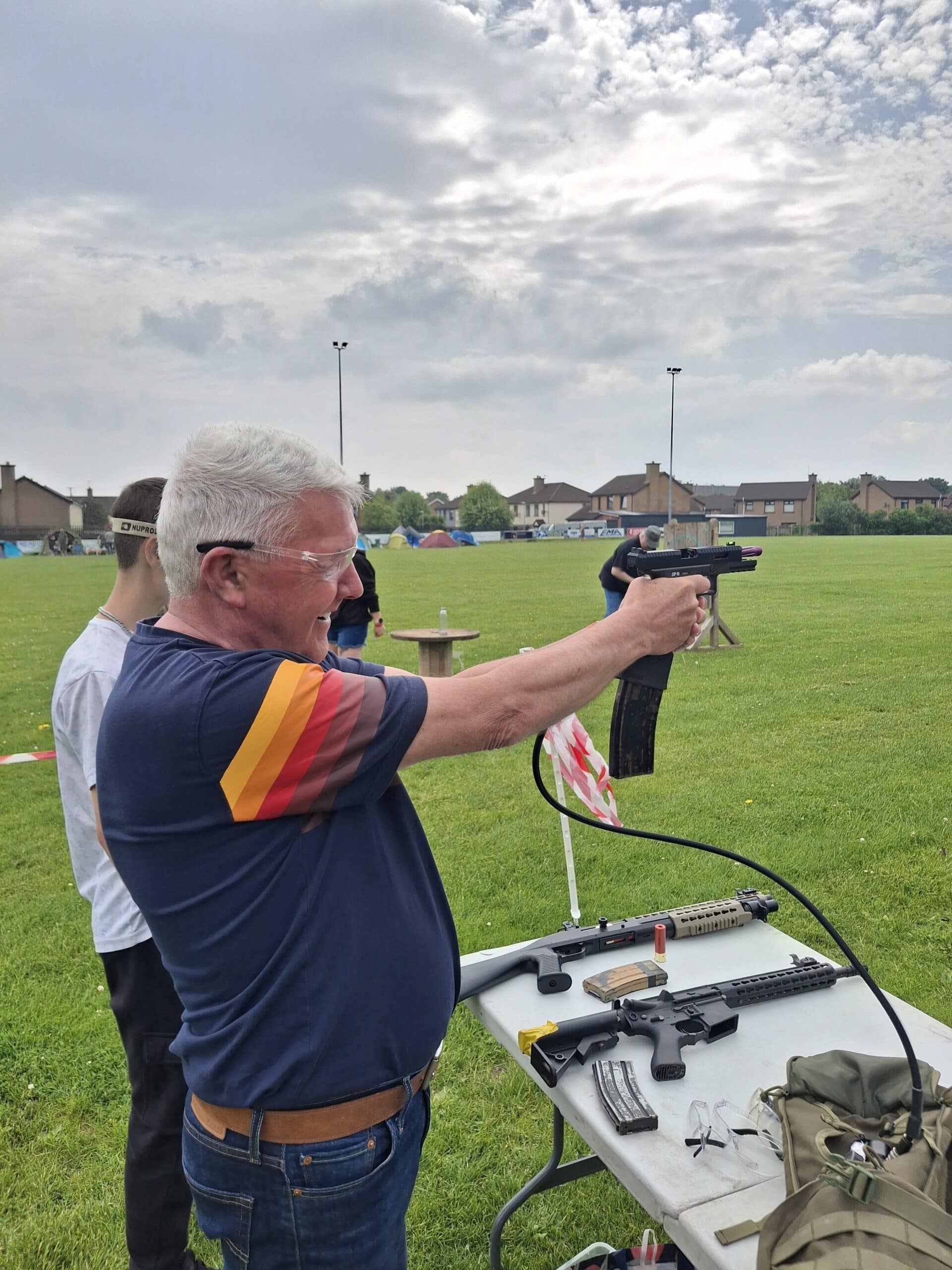 Man in safety goggles aiming an airsoft gun at a shooting range outdoors.