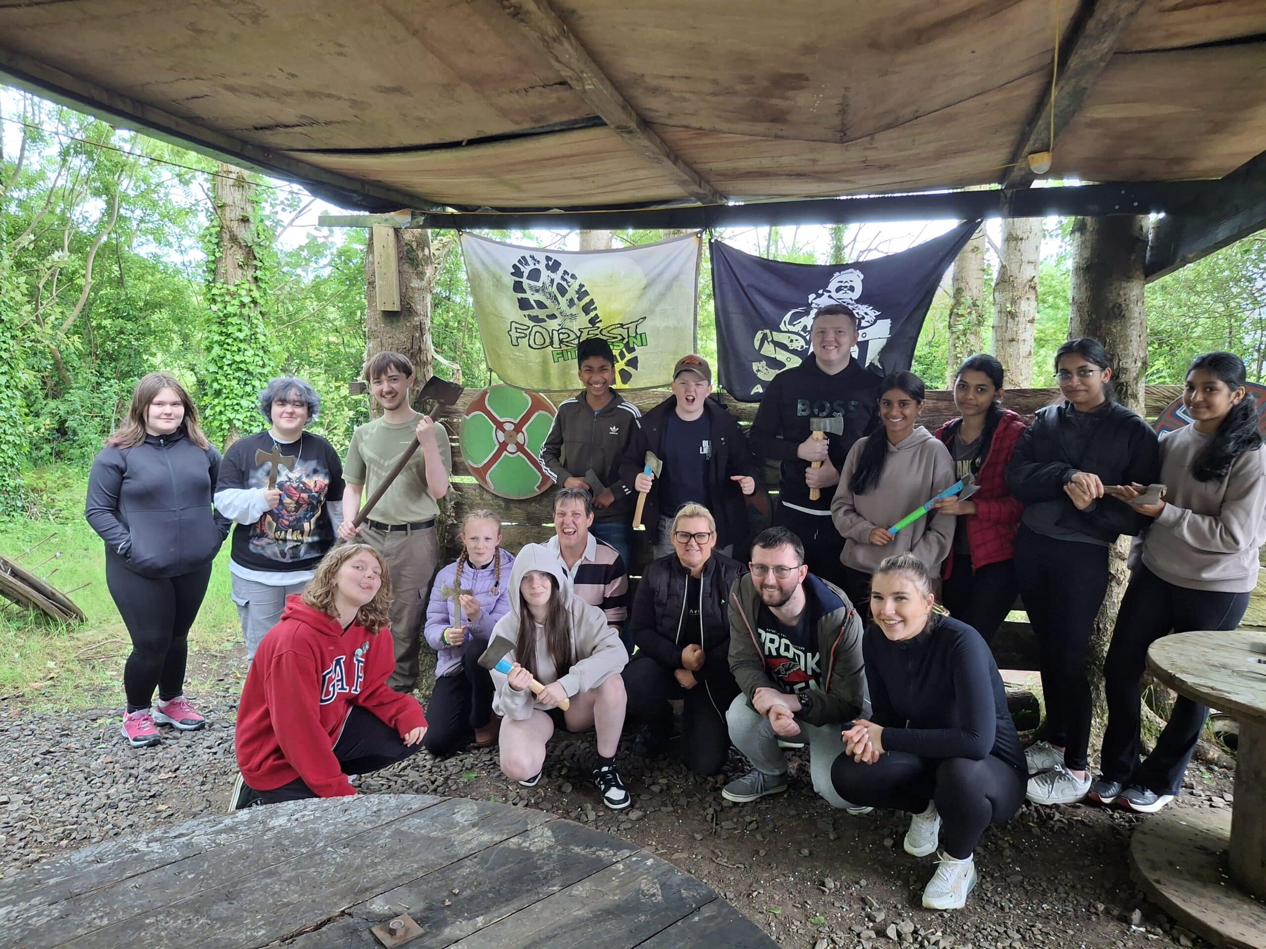 Group of people posing with axes under a canopy in a forest setting, with banners in the background.