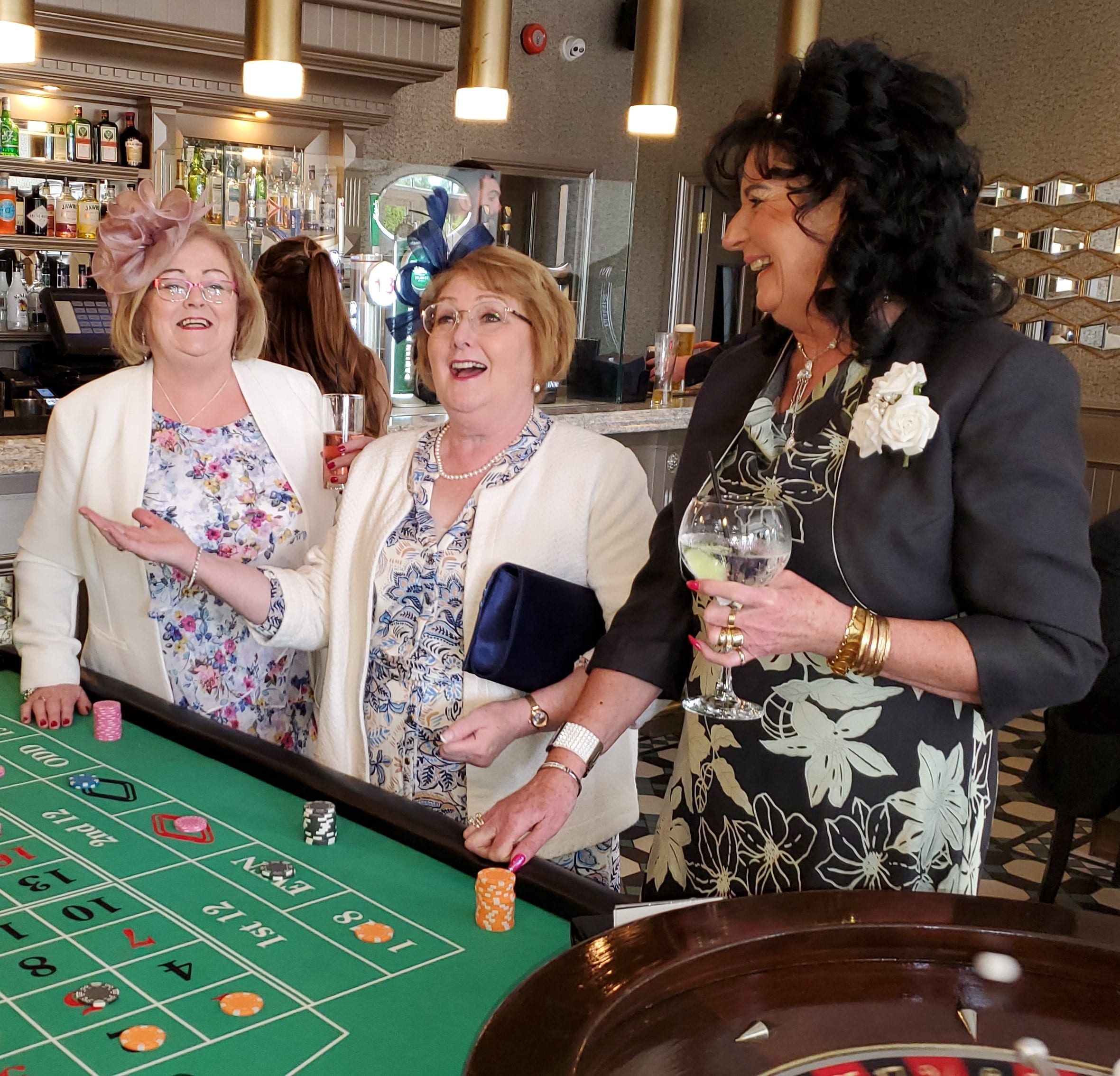 Three women dressed elegantly enjoying a game of roulette at a casino, with drinks in hand.
