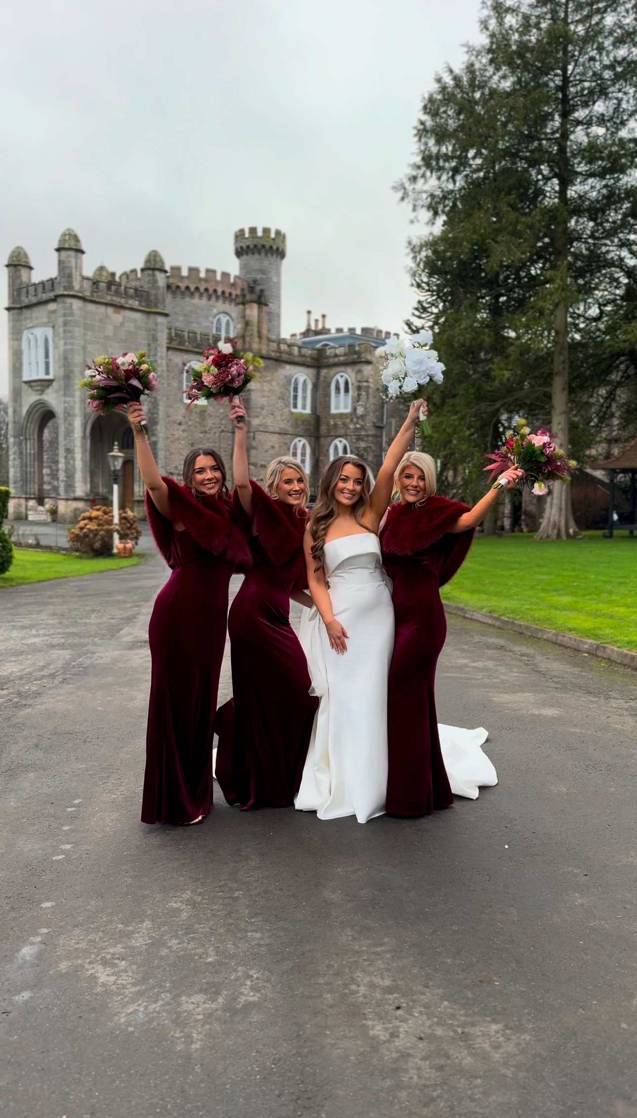 Bride and bridesmaids in front of a castle, holding bouquets, wearing burgundy dresses and a white wedding gown.