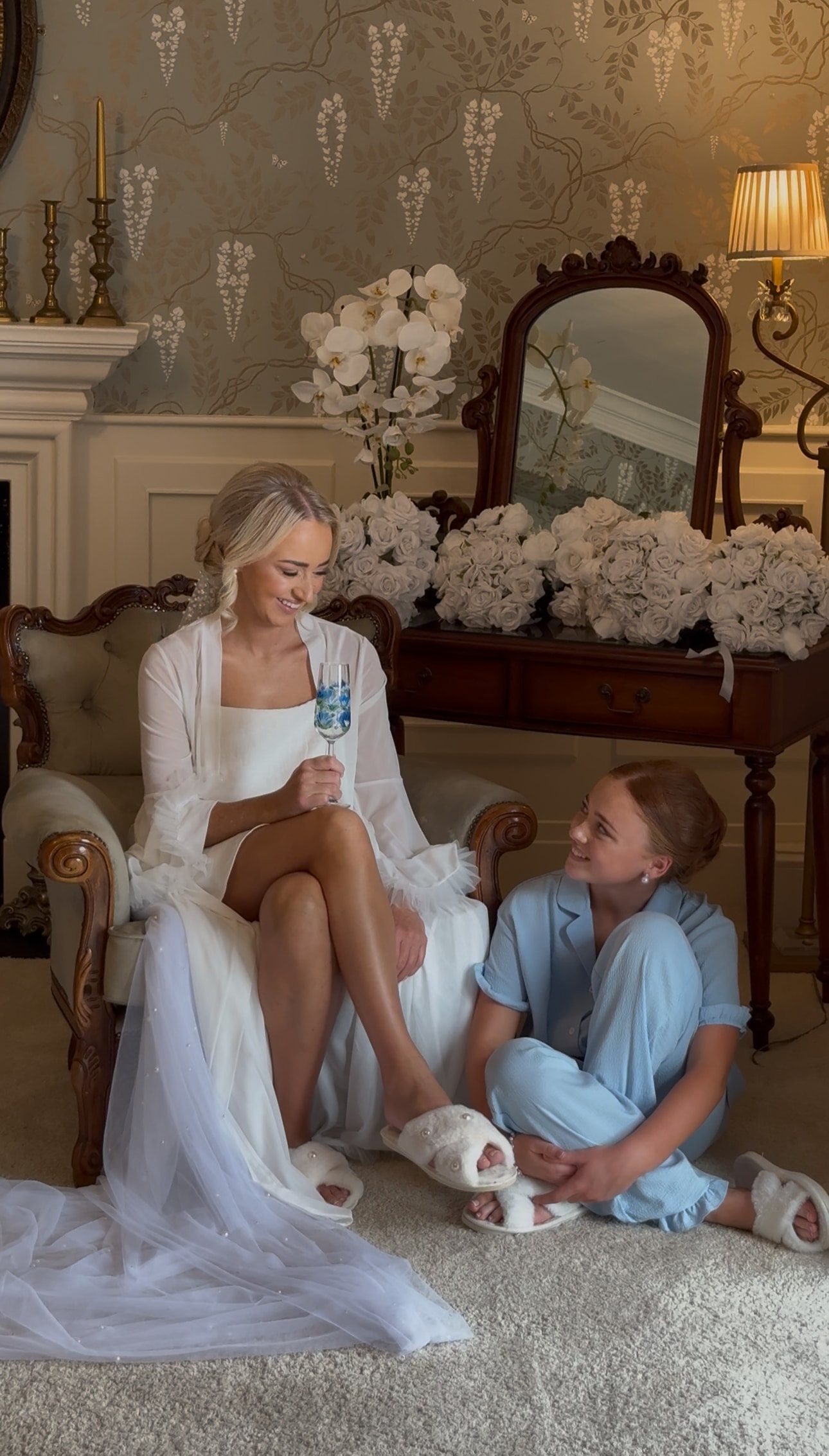 Bride sitting with a young girl, both in pajamas, in a vintage room with floral decor.
