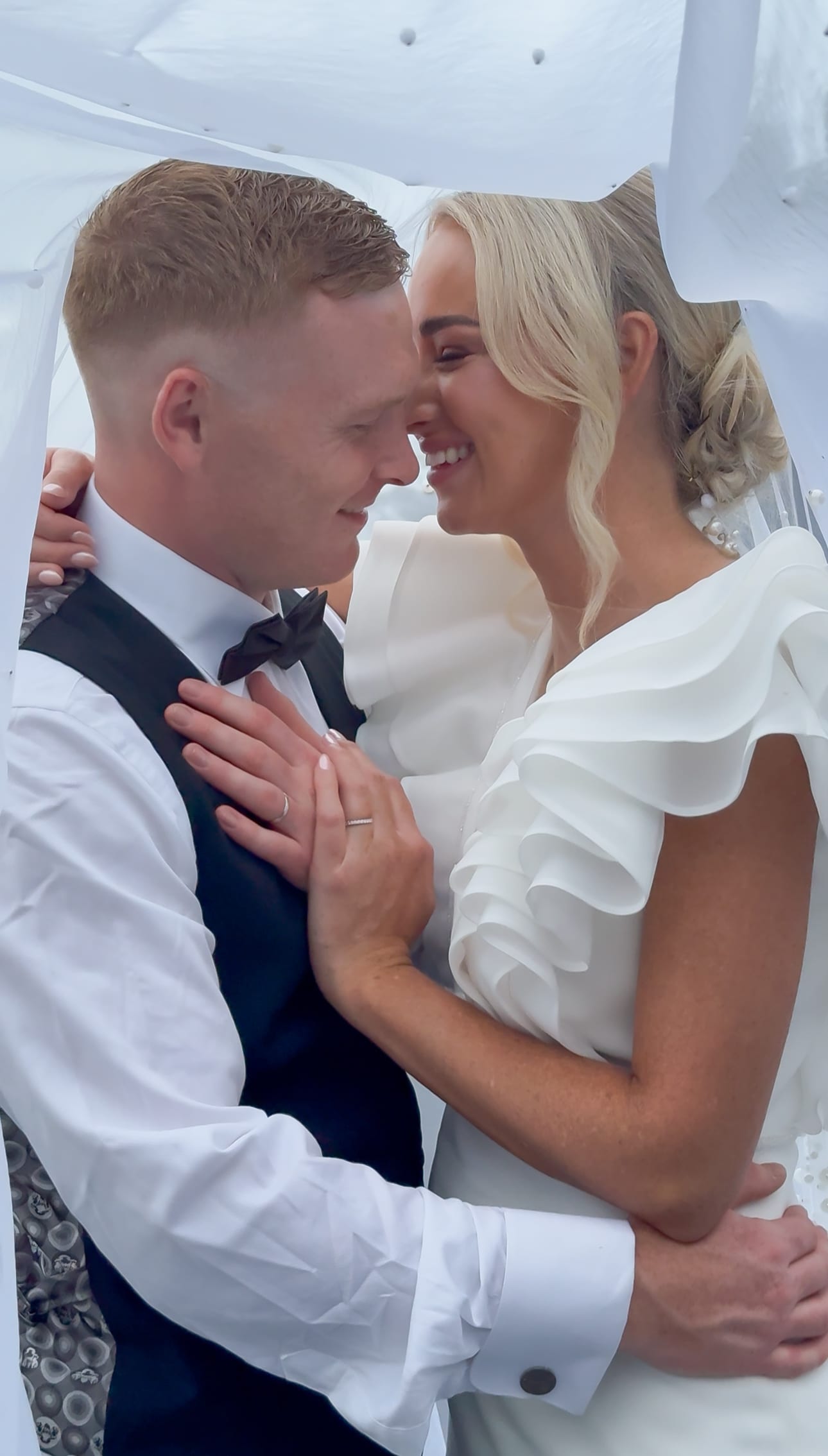 Bride and groom embracing under veil, smiling at each other on wedding day.