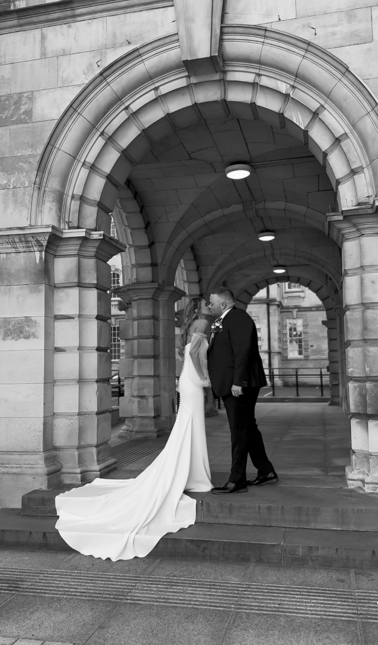 Bride and groom kiss under an arched stone doorway, captured in black and white.