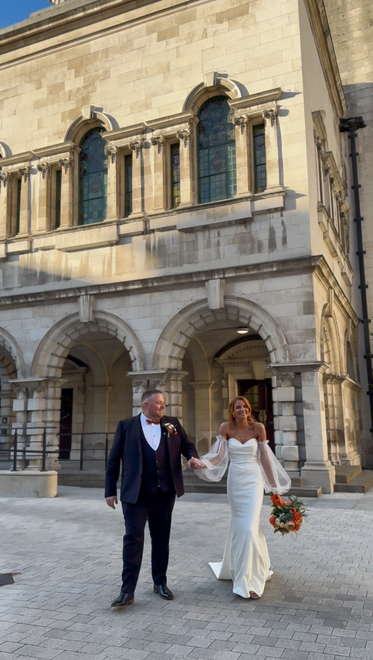 Bride and groom hold hands outside a historic stone building, the bride holding a bouquet.