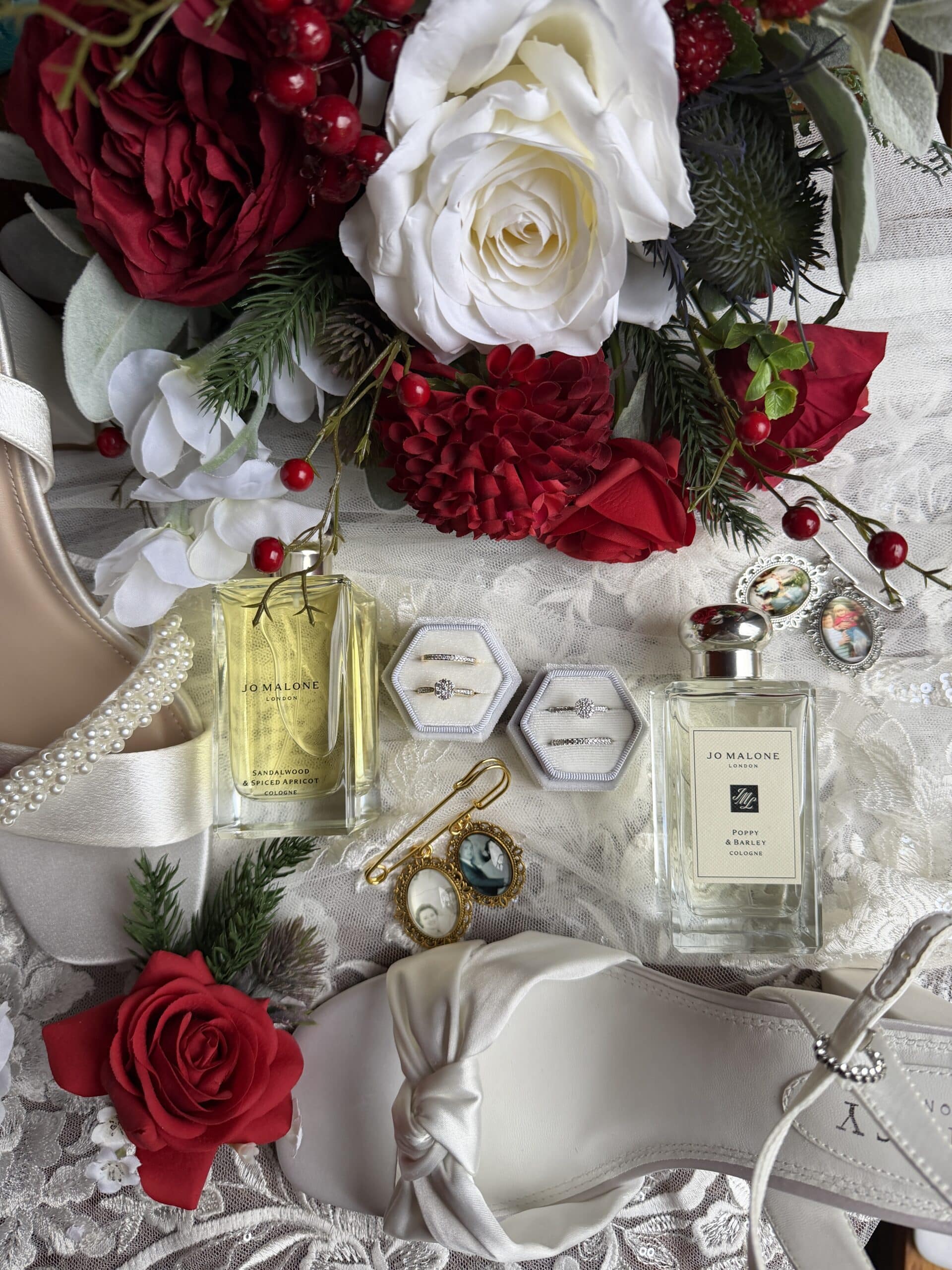 Floral wedding arrangement with red and white roses, perfume bottles, jewelry, and white satin shoes.