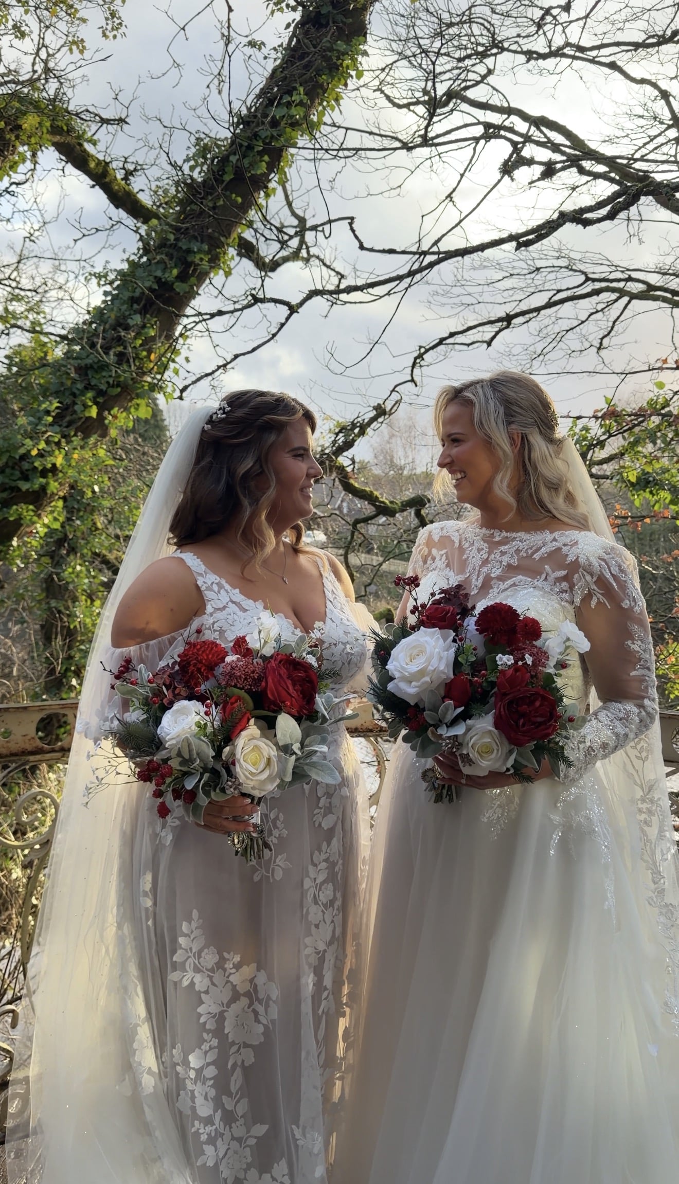 Two brides in lace wedding dresses holding bouquets with red and white flowers, standing outdoors in front of trees.