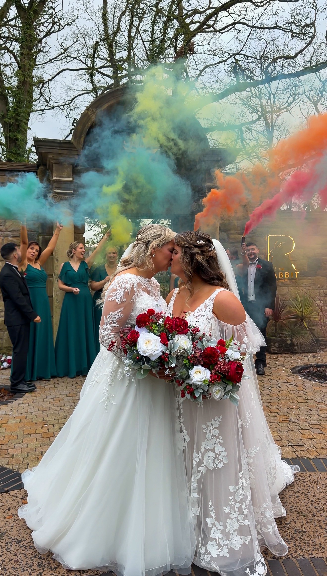 Two brides kissing with vibrant colored smoke in the background, surrounded by wedding party in green dresses.