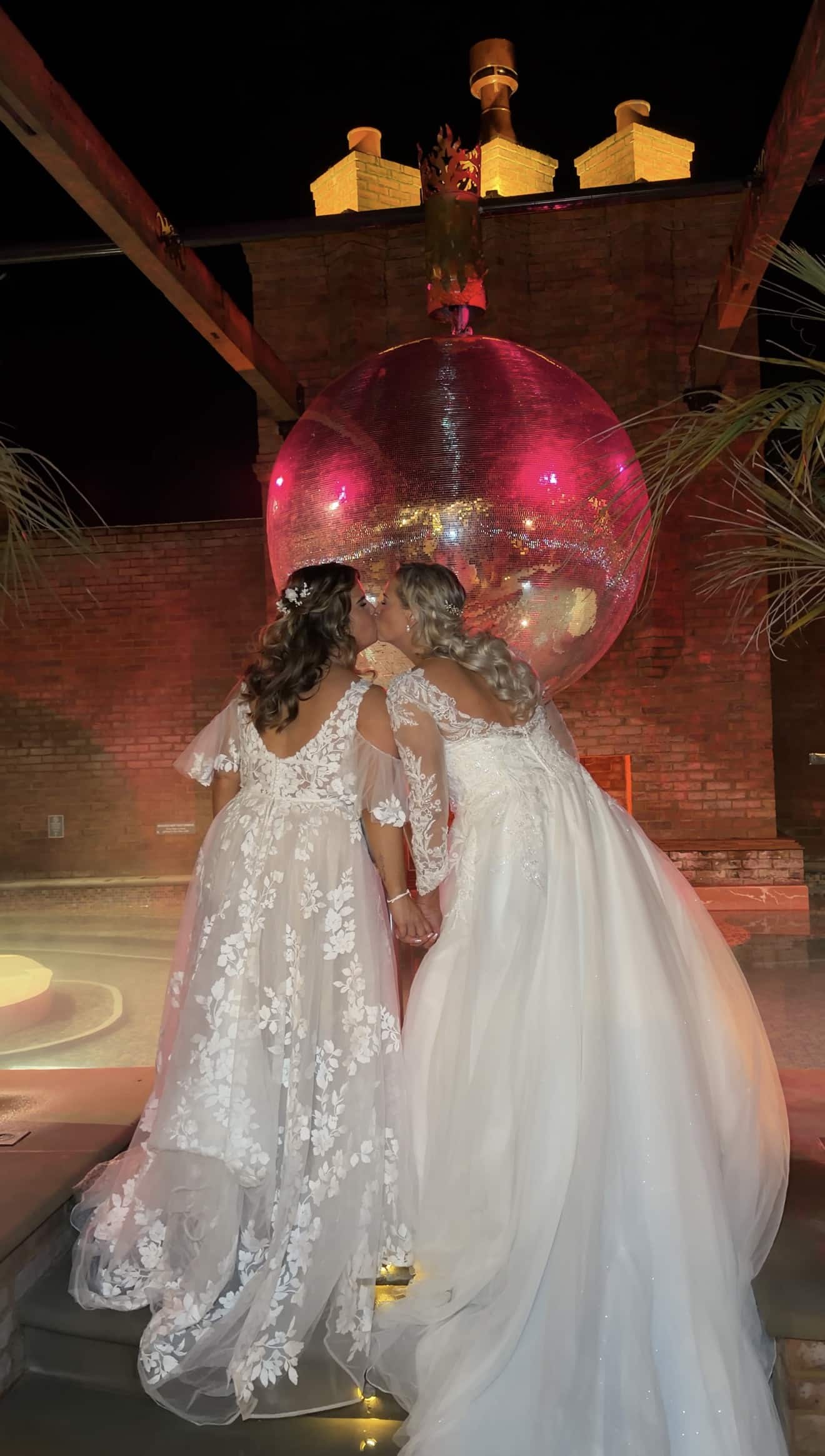 Two brides in elegant gowns share a kiss under a large glowing disco ball, surrounded by brick walls and palm leaves.