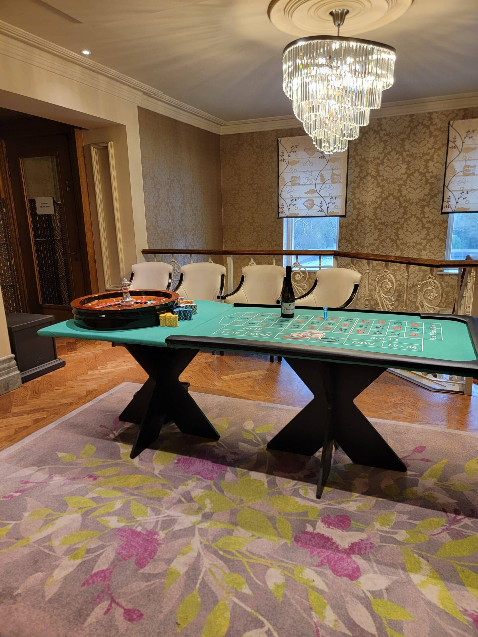 Luxurious room with a roulette table under an elegant chandelier, featuring ornate wallpaper and plush floral carpet.